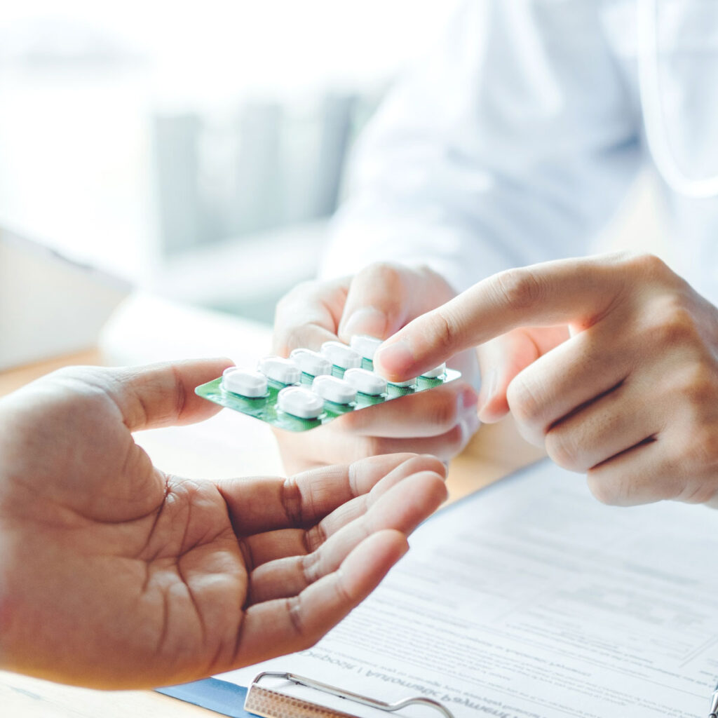A doctor hands a blister pack of white pills to a patient. The patient’s hand is reaching out to receive the medication. A clipboard with paperwork is visible on the table.