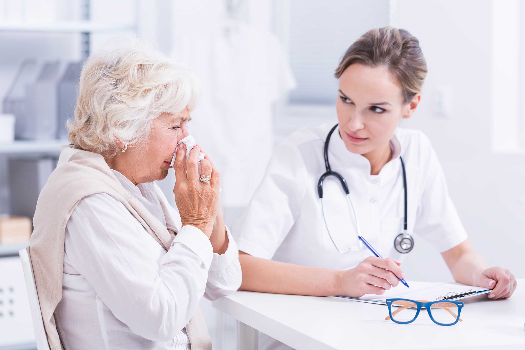 An older woman blows her nose with a tissue while sitting across from a doctor, who is taking notes and listening attentively in a bright medical office.