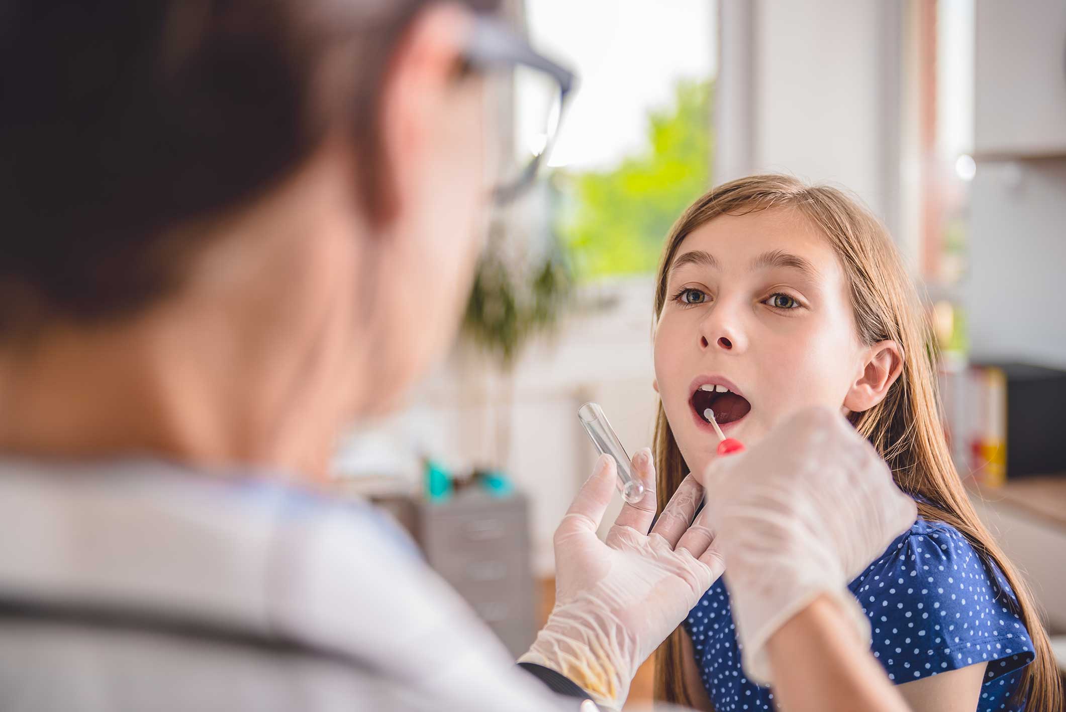 A healthcare professional wearing gloves swabs the inside of a young girls mouth during a medical examination in a brightly lit room.