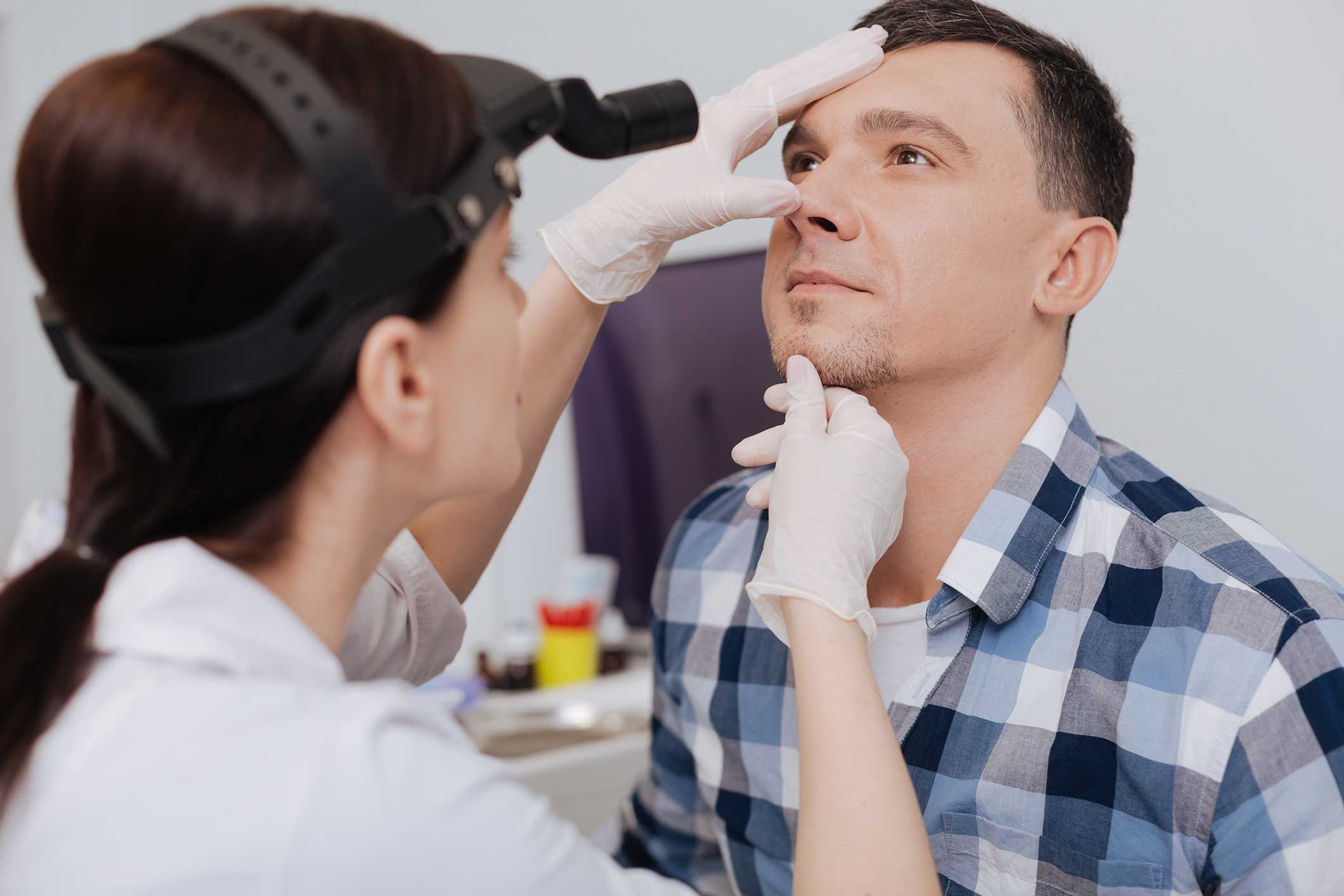 A doctor wearing gloves and a headlamp examines a man’s nose and face. The man, in a blue plaid shirt, looks upward. The clinical setting includes medical tools and supplies in the background.