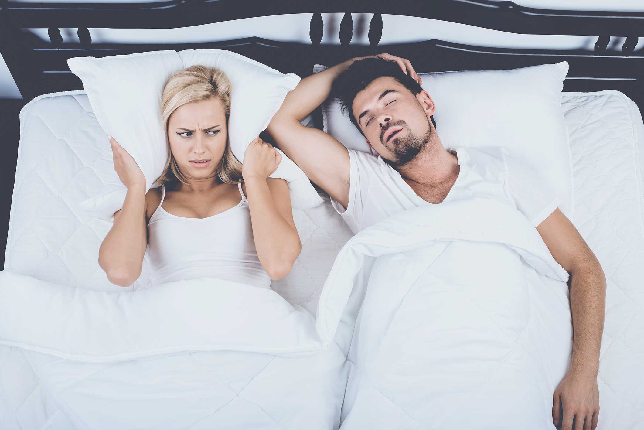 A woman looks annoyed while covering her ears with a pillow as a man sleeps beside her, appearing to snore, in a bed with white sheets.