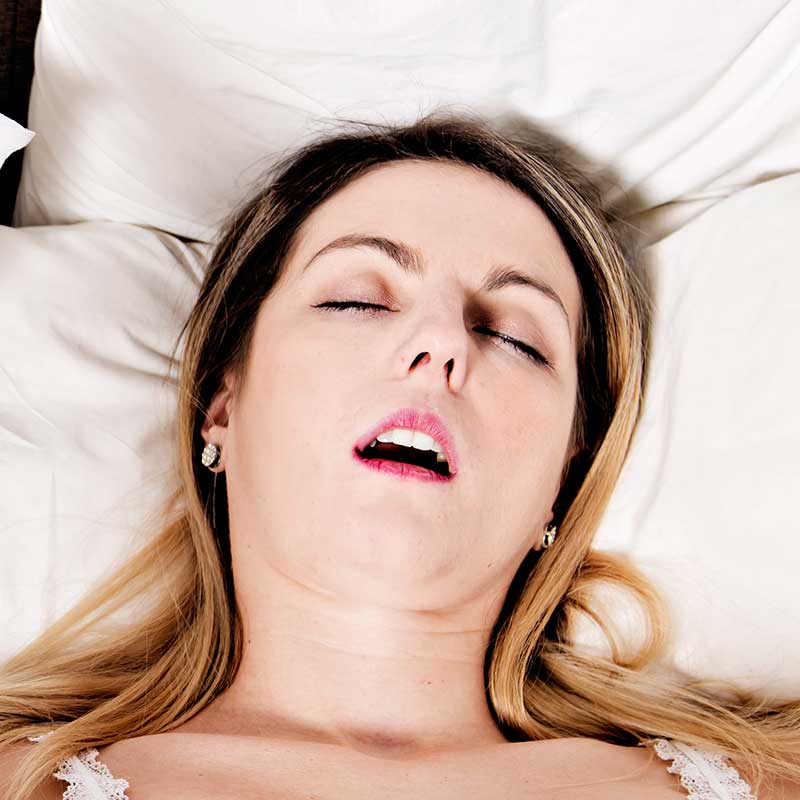 A woman with light hair lies on a bed with her eyes closed and mouth slightly open, appearing to be asleep or resting.