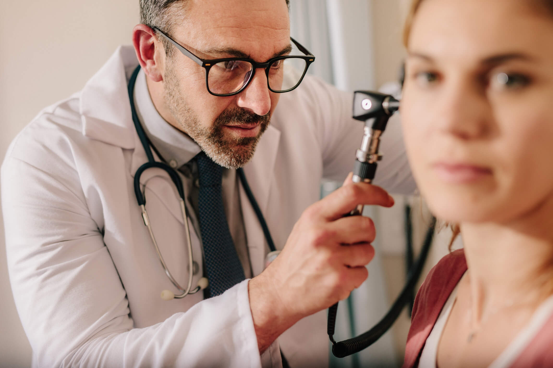 A doctor wearing glasses examines a womans ear with an otoscope. The doctor appears focused, while the woman looks ahead, slightly out of focus. Both are indoors in a clinical setting.