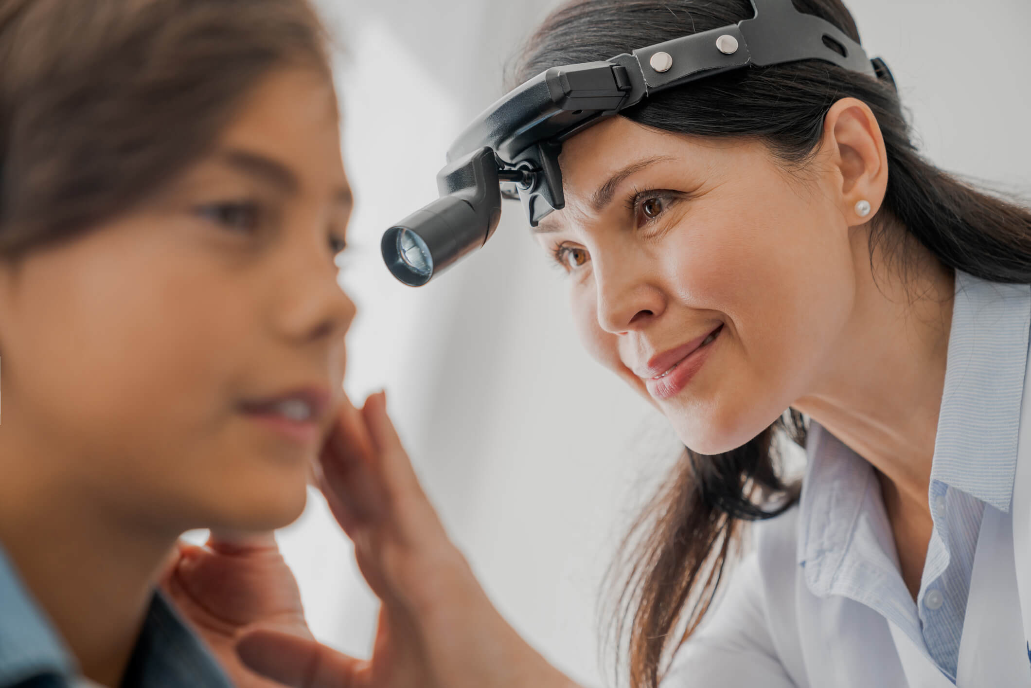 A doctor wearing a headlamp examines a young patient’s face closely, smiling gently and touching the patient’s cheek with one hand during a medical check-up.