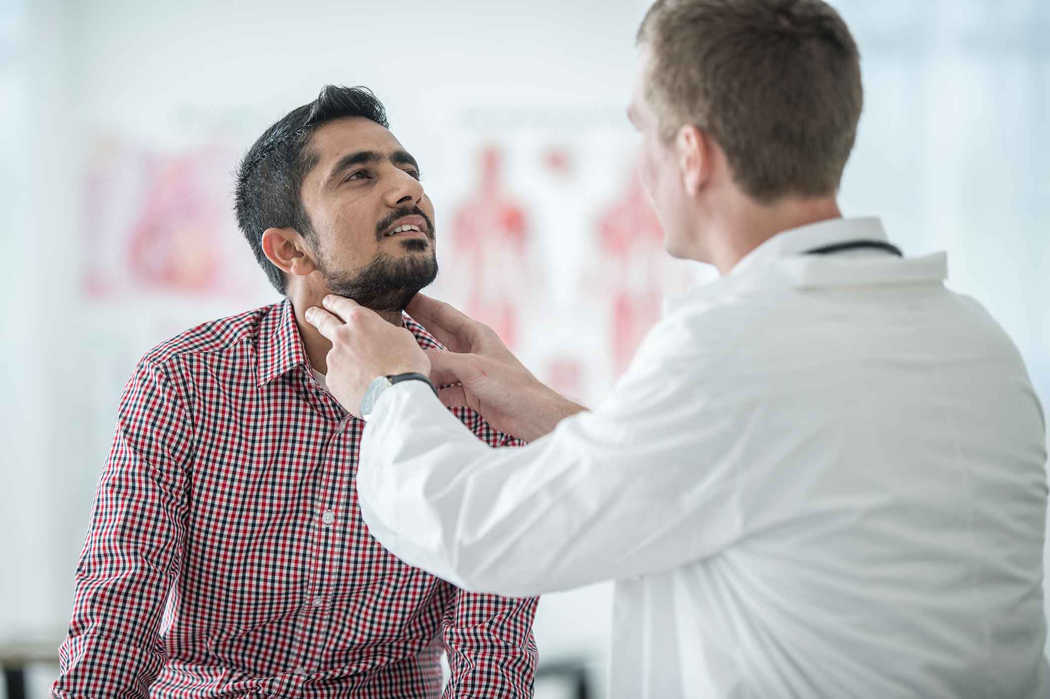 A doctor in a white coat examines a mans neck and throat while the man sits on an exam table and looks upward. The setting appears to be a medical office.