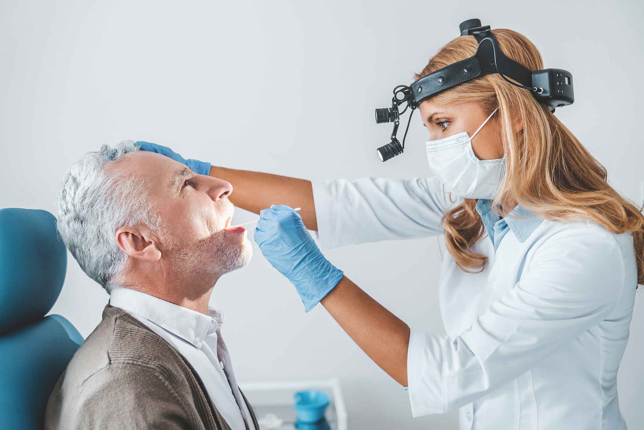 A female doctor wearing a mask and headlamp examines an older male patient’s mouth with a swab. The patient is seated, and both are in a medical office.
