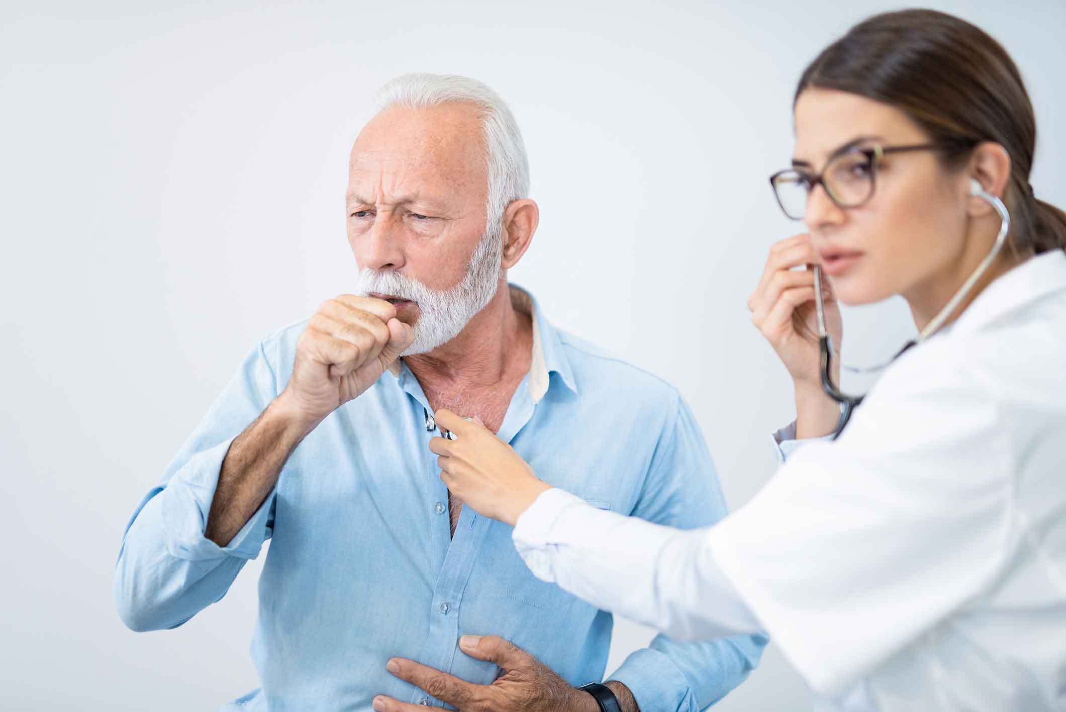 An older man coughs into his hand while a doctor with a stethoscope listens to his chest, both appearing serious, in a medical examination setting.