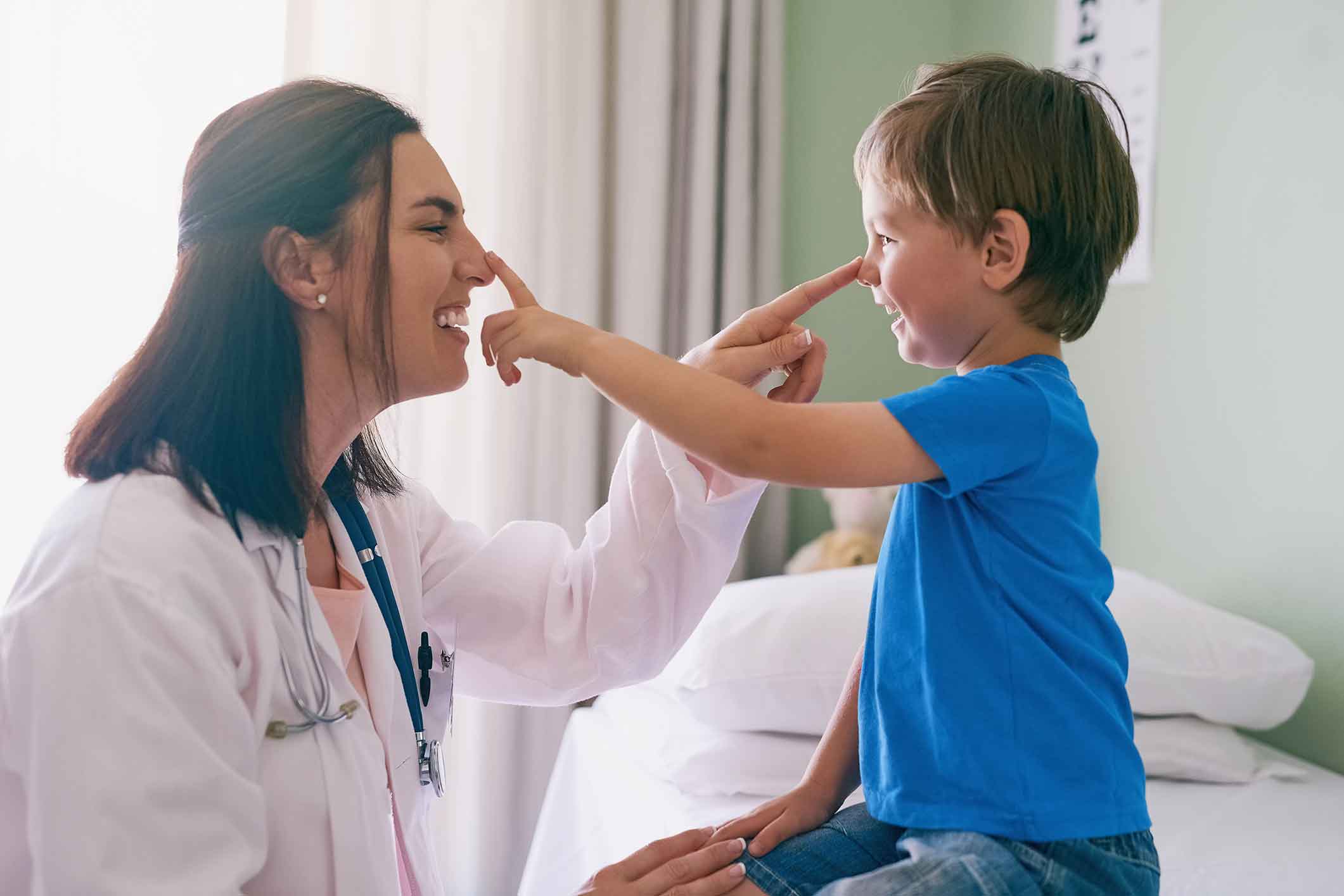 A smiling doctor and a young boy touch each other’s noses while sitting in a medical office, both laughing and enjoying the playful moment.