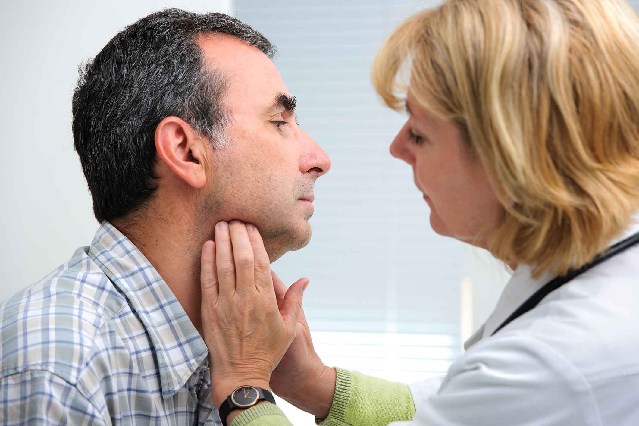 A doctor examines a mans neck by gently pressing below his jaw, possibly checking his lymph nodes or thyroid. Both appear focused and calm during the medical checkup.
