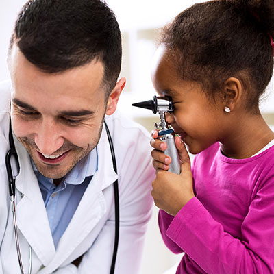 A smiling doctor leans toward a young girl in a pink shirt who playfully examines his ear with an otoscope.