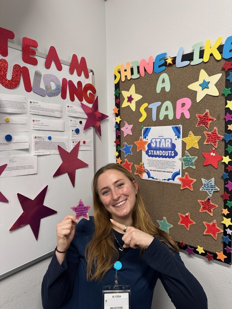 A woman smiles and points at a colorful bulletin board covered with stars and the phrase SHINE LIKE A STAR next to a “TEAM BUILDING” whiteboard. She wears a name badge and stands in a cheerful, decorated room.