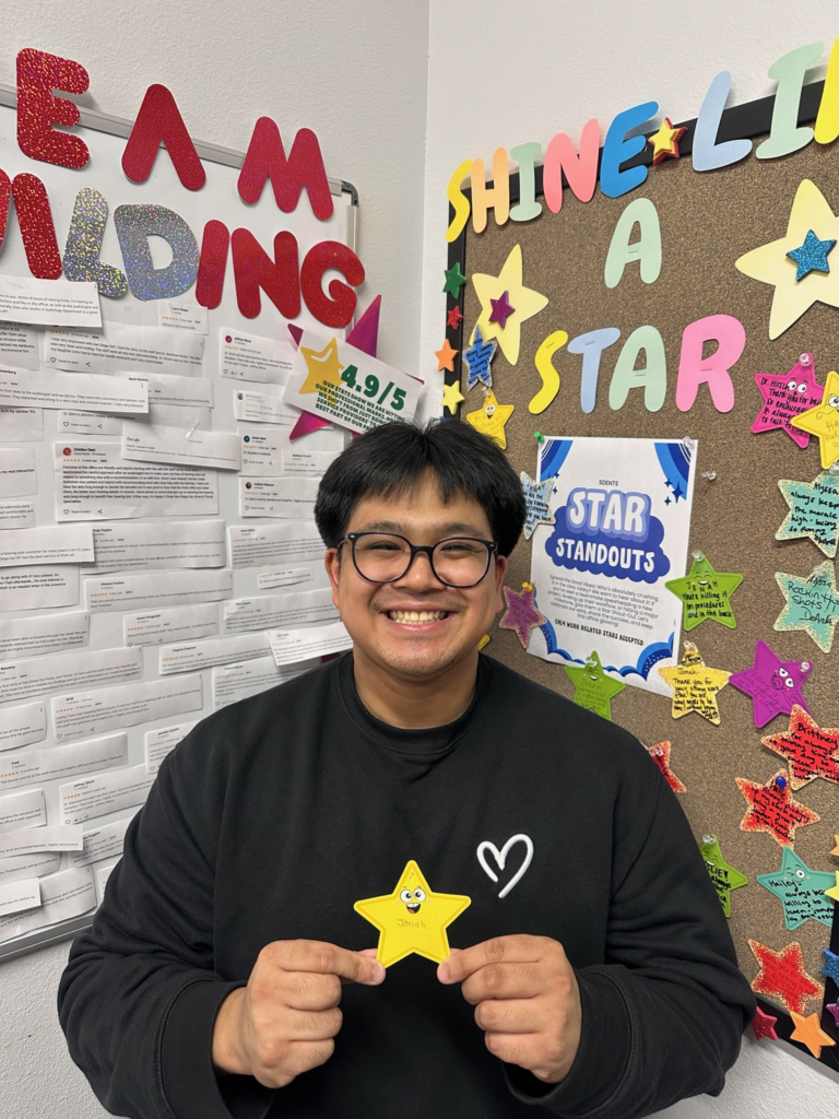 A person smiling and holding a yellow star stands in front of colorful bulletin boards decorated with stars and positive messages like SHINE LIKE A STAR and TEAM BUILDING.