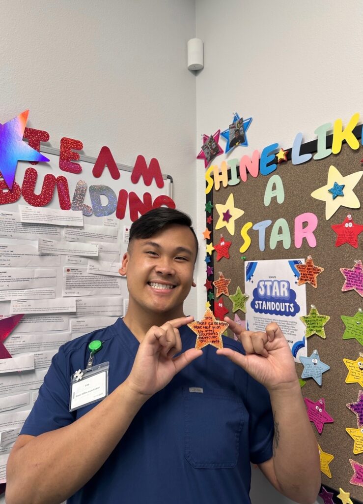 A smiling person in blue scrubs holds a star-shaped paper in front of a bulletin board decorated with colorful stars and the words “SHINE LIKE A STAR.” Nearby is another board labeled “TEAM BUILDING.”.