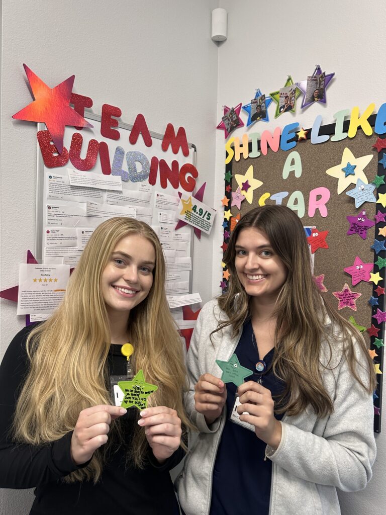 Two women stand smiling, holding paper stars. Behind them are colorful bulletin boards with stars and the phrases TEAM BUILDING and SHINE LIKE A STAR. The setting appears to be an office or classroom.