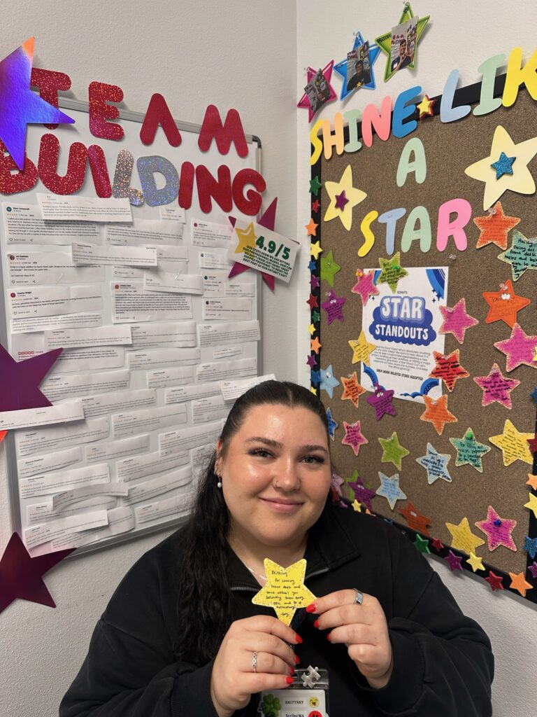 A woman smiles, holding a yellow star-shaped note. Behind her are bulletin boards covered with colorful stars, notes, and letters spelling TEAM BUILDING and SHINE LIKE A STAR.