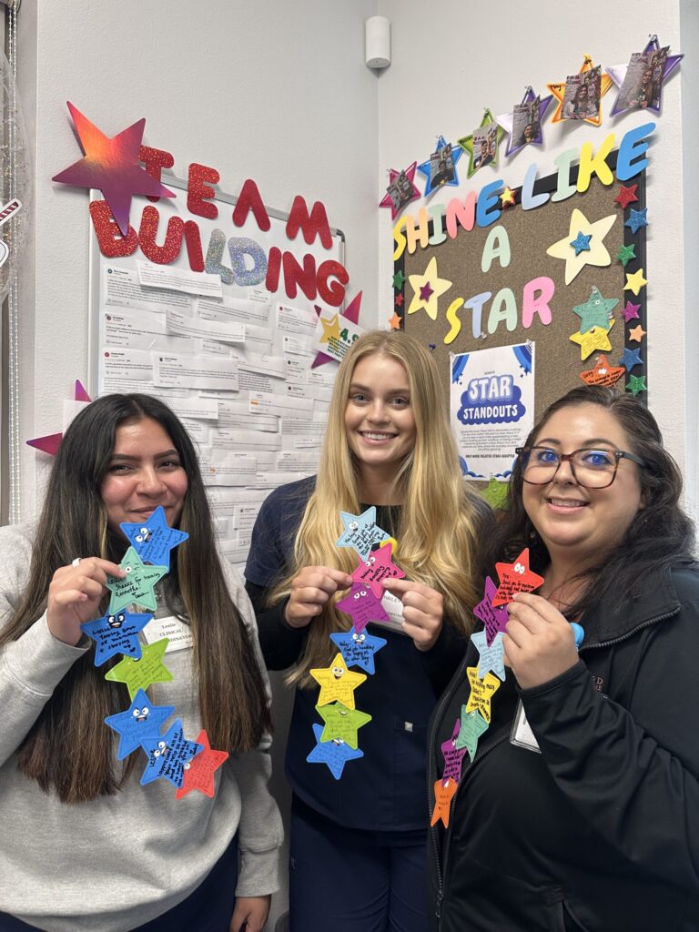 Three women smile and hold colorful paper stars with messages in an office decorated with stars and signs reading “Team Building” and “Shine Like a Star.” A corkboard is covered in more star-shaped notes.