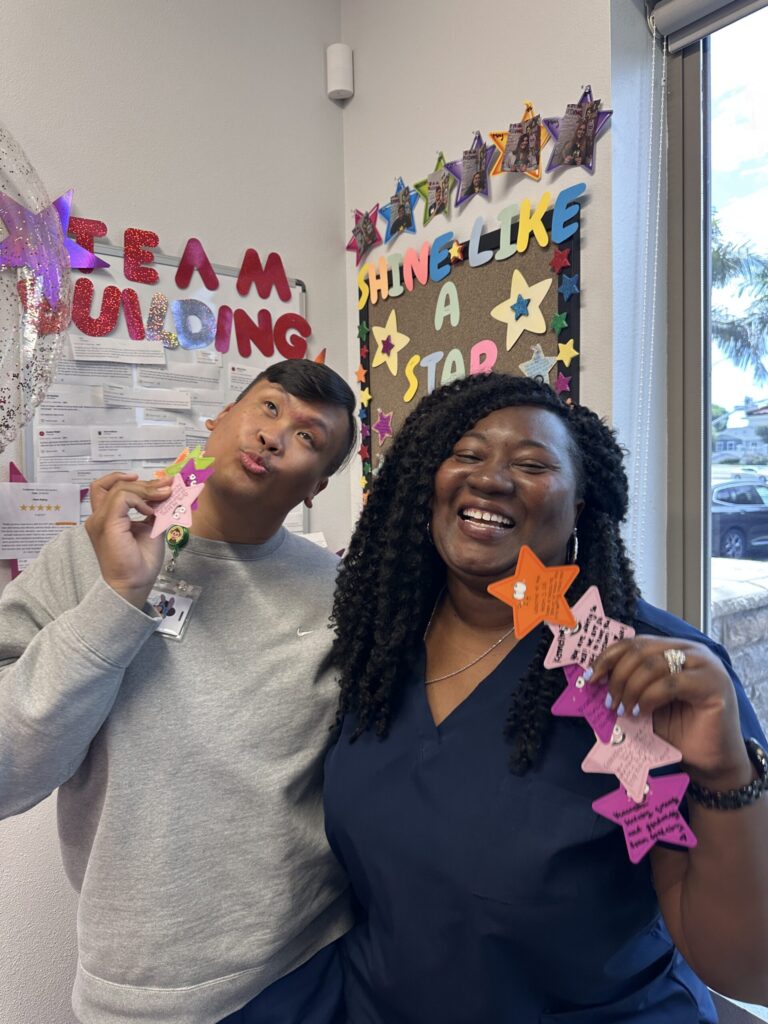 Two people smile and hold colorful star-shaped notes in a decorated room with “SHINE LIKE A STAR” and “TEAM BUILDING” signs on the wall behind them.