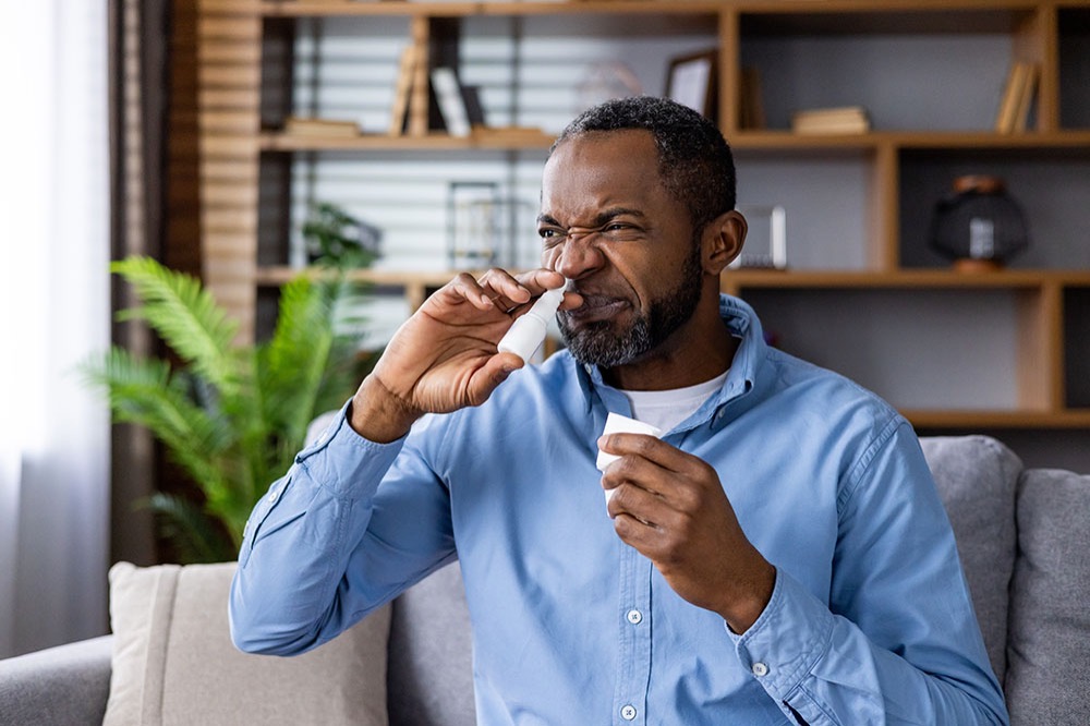 A man sitting on a couch squirts nasal spray into his nose, holding the bottle in one hand and closing his eyes with a slight grimace. A plant and shelves are visible in the background.