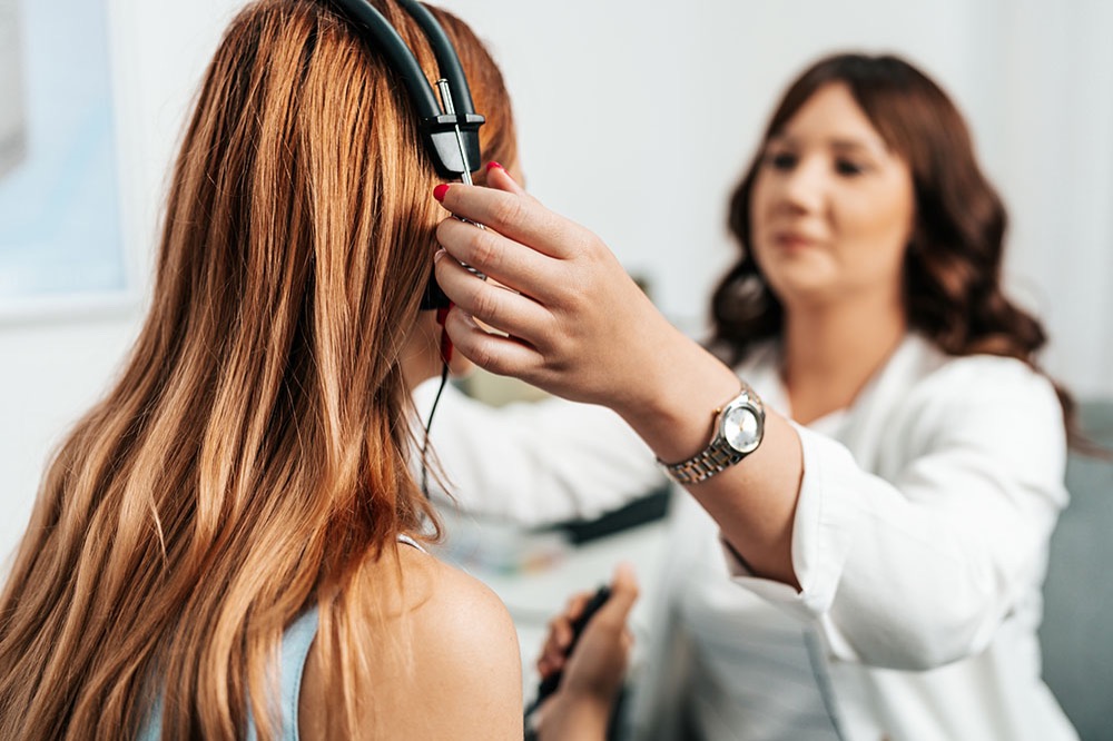 A woman in a white coat places headphones on another womans head, likely preparing her for a hearing test in a medical or audiology office.