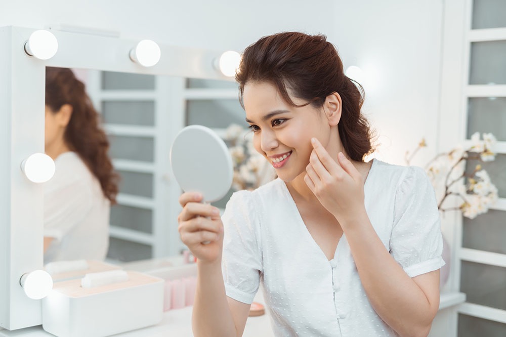 A woman in a white top sits at a vanity table with lights, holding a small round mirror and touching her cheek while smiling. The background includes decorative flowers and beauty products.