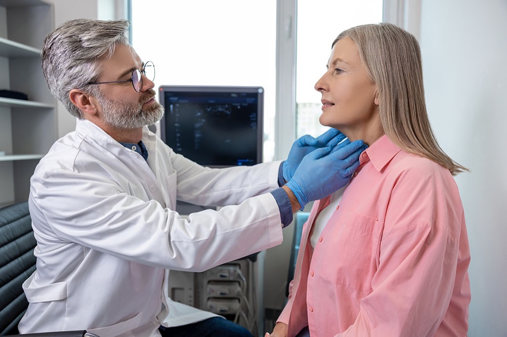 A doctor wearing gloves examines a woman’s neck in a medical office, possibly checking her thyroid. The woman is seated and wearing a pink shirt. Medical equipment and a window are visible in the background.
