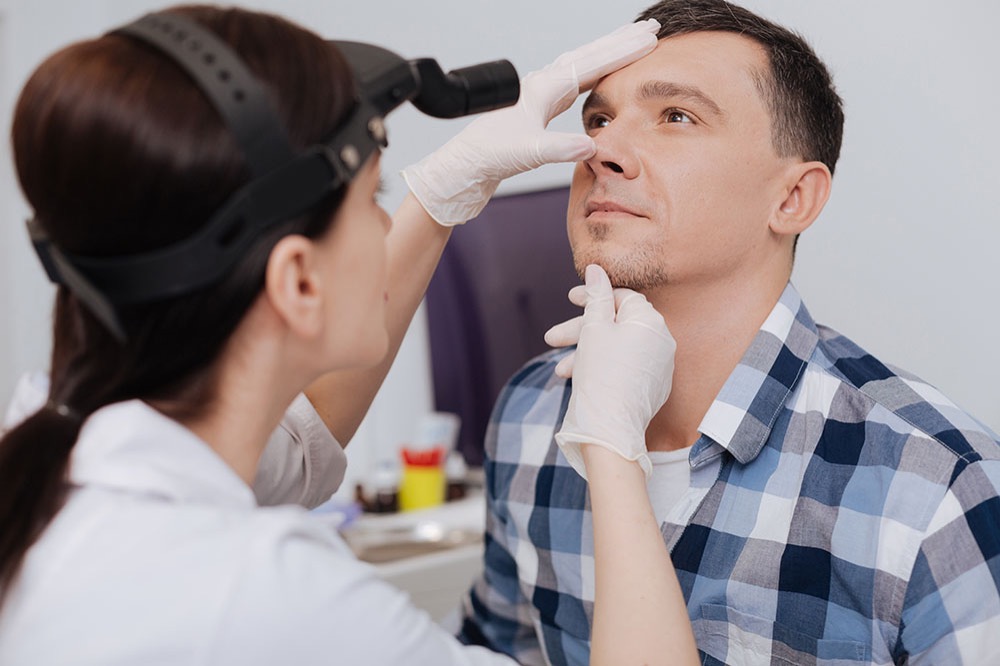 A doctor wearing gloves and a headlamp examines a mans nose and throat. The man, in a blue plaid shirt, looks upward while the doctor gently touches his face in a clinical setting.