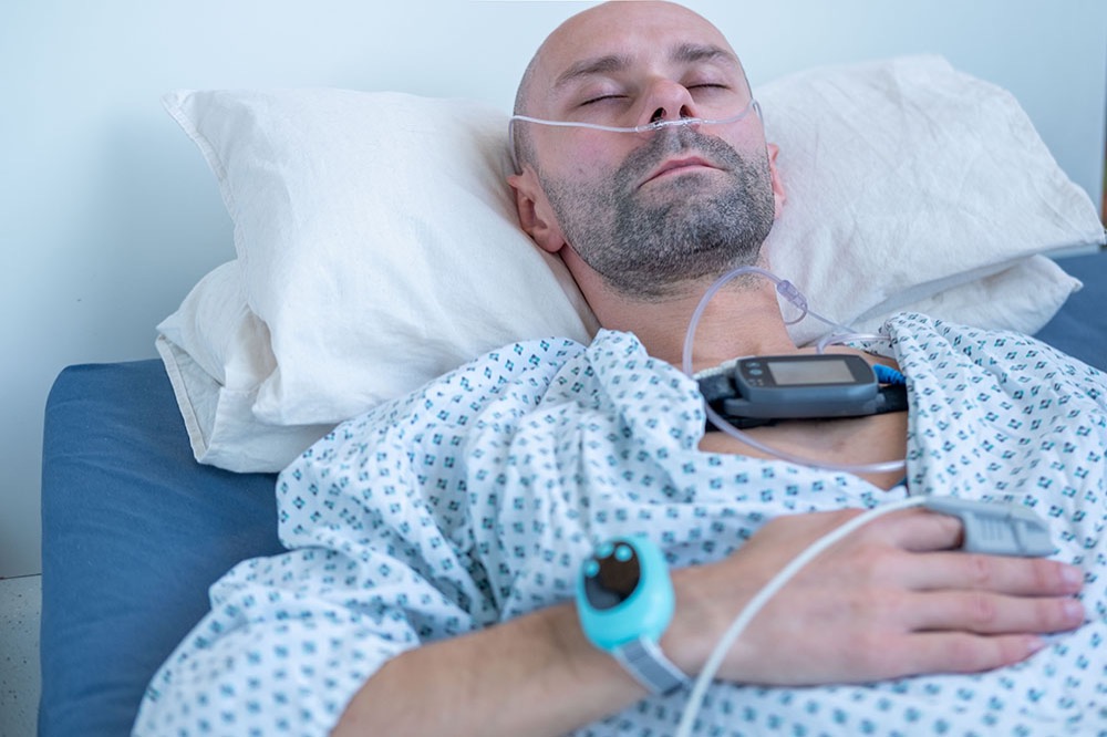 A man with a nasal oxygen tube lies in a hospital bed, eyes closed. He wears a hospital gown, a medical device on his chest, a finger monitor, and a blue wristband.