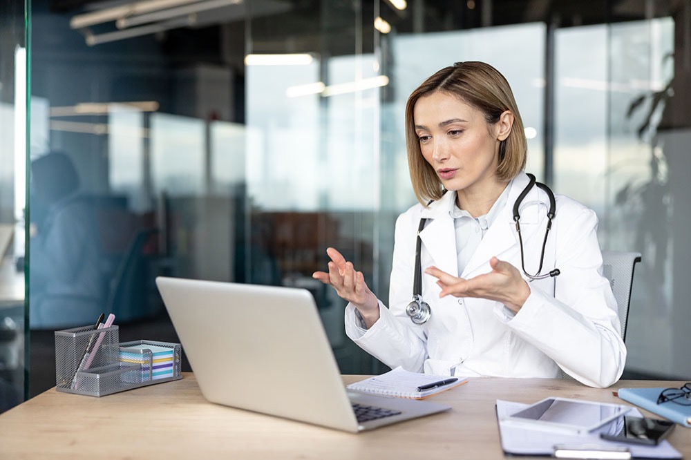 A doctor wearing a white coat and stethoscope sits at a desk, gesturing while speaking during a video call on a laptop in a modern office setting.