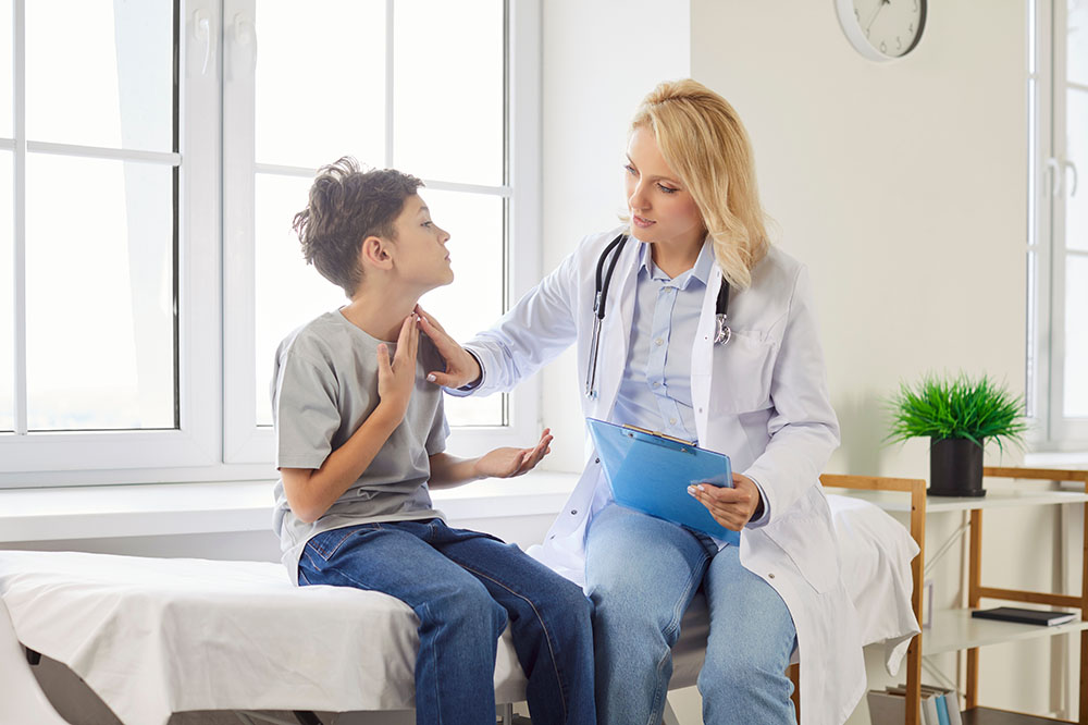 A doctor with a clipboard examines a boy sitting on an exam table, gently touching his neck. They appear to be discussing his symptoms in a bright medical office.